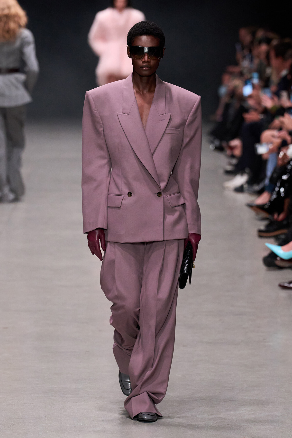 Model walks the runway in an oversized dusty-pink double-breasted suit with wide shoulders, burgundy gloves, sunglasses, and a black clutch.