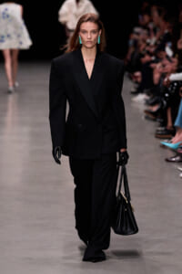 Model walking the runway in an oversized black blazer and wide-leg pants, black gloves, turquoise earrings, and a black handbag.