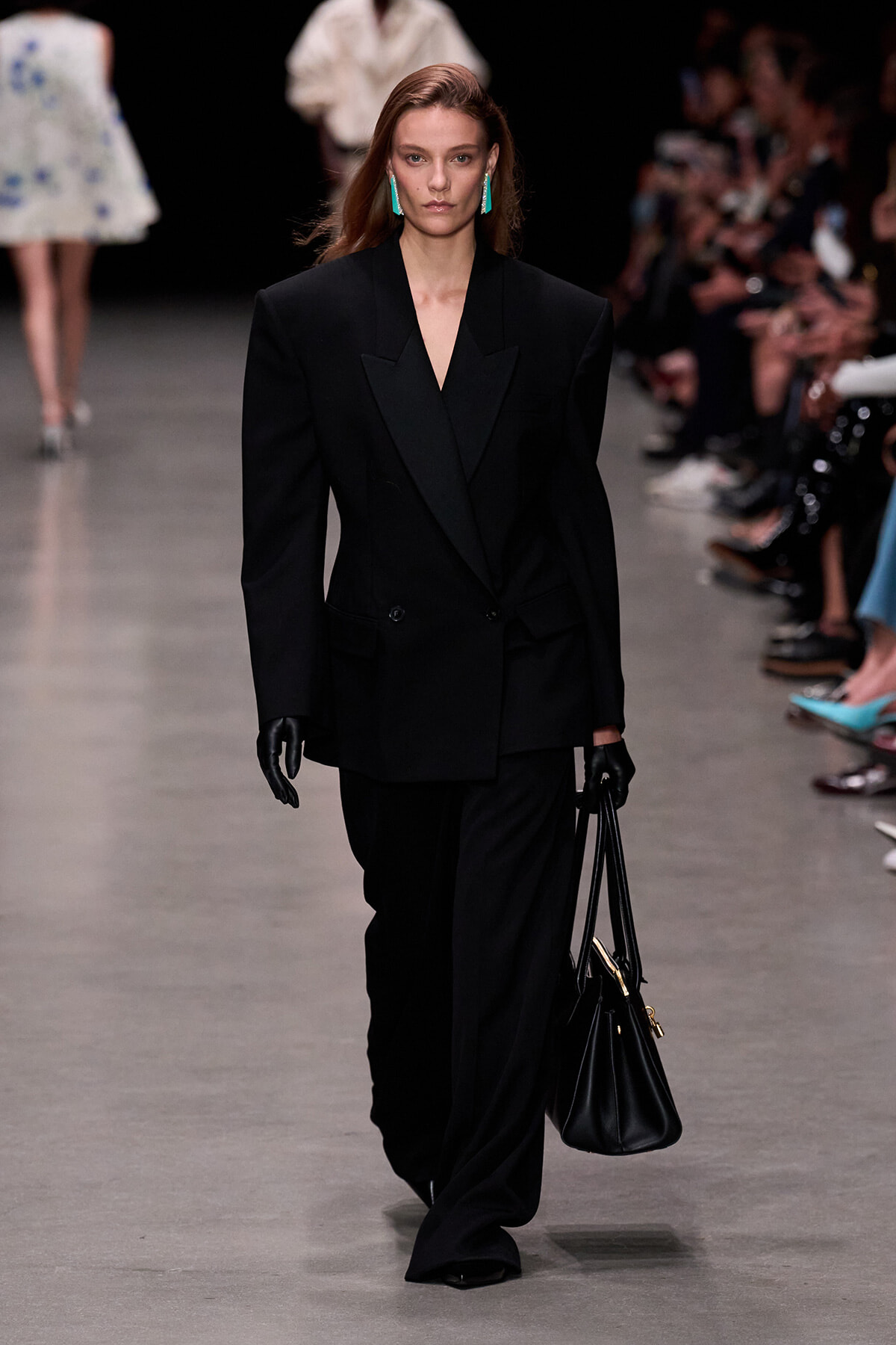 Model walking the runway in an oversized black blazer and wide-leg pants, black gloves, turquoise earrings, and a black handbag.