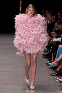 Model on a runway wearing a voluminous light-pink fringe dress, with spectators along the sides.