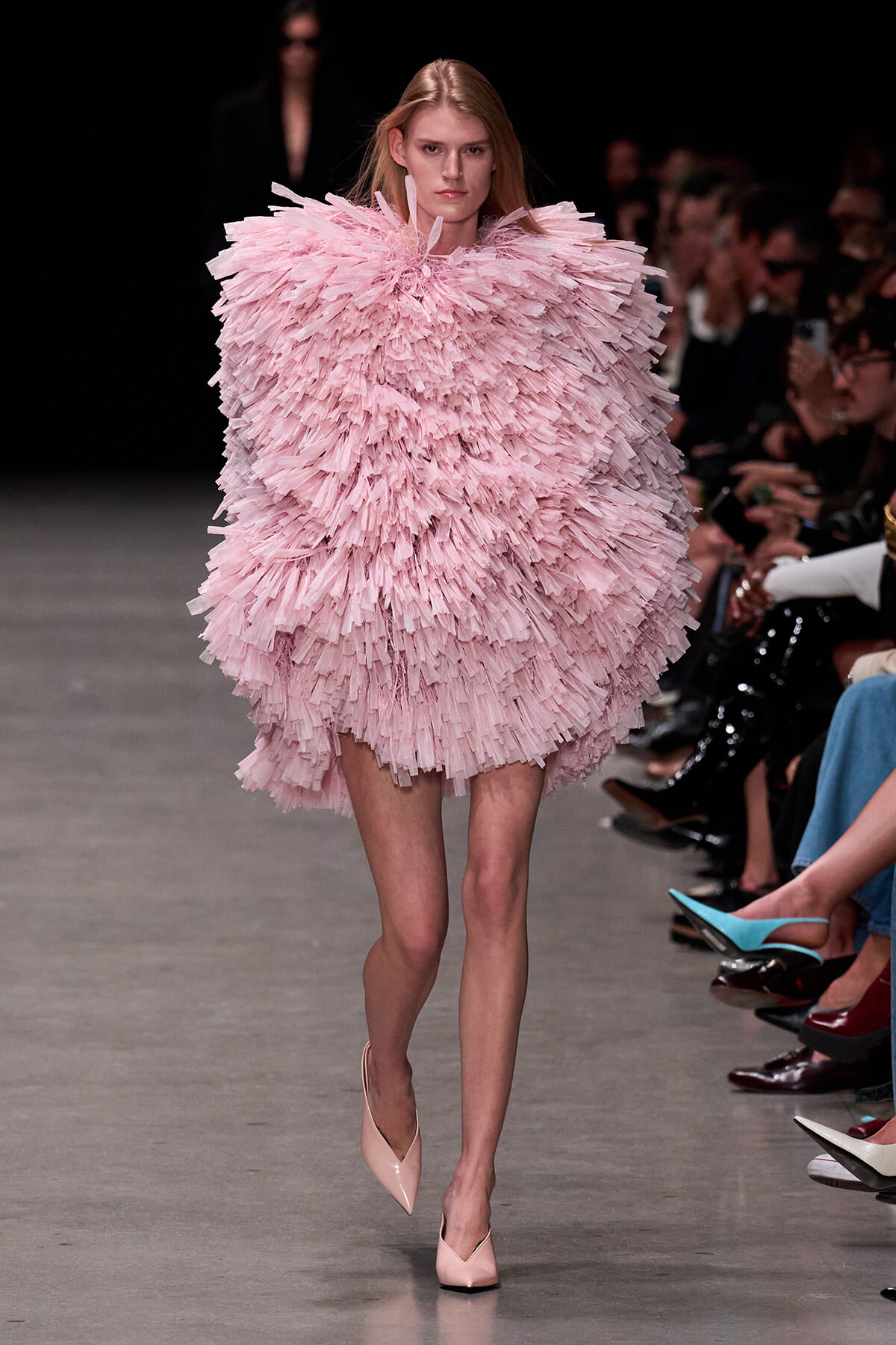 Model on a runway wearing a voluminous light-pink fringe dress, with spectators along the sides.