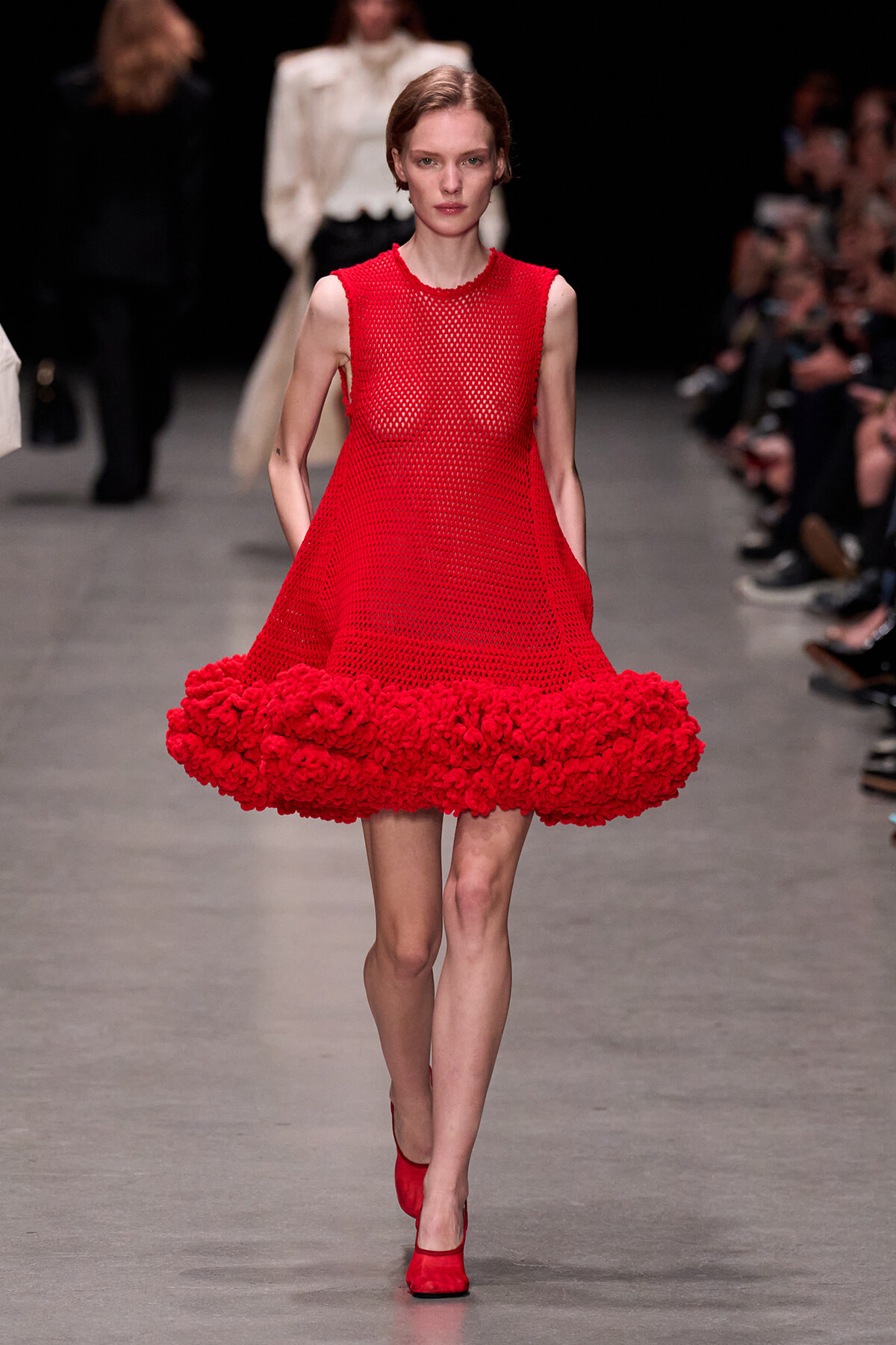 Model walks the runway in a red mesh sleeveless dress with a voluminous pom-pom hem, paired with red heels.
