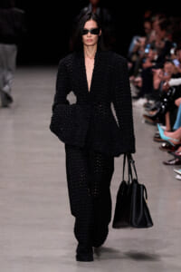 Model walks the runway in an all-black textured suit with oversized jacket, black sunglasses, and a black tote bag.