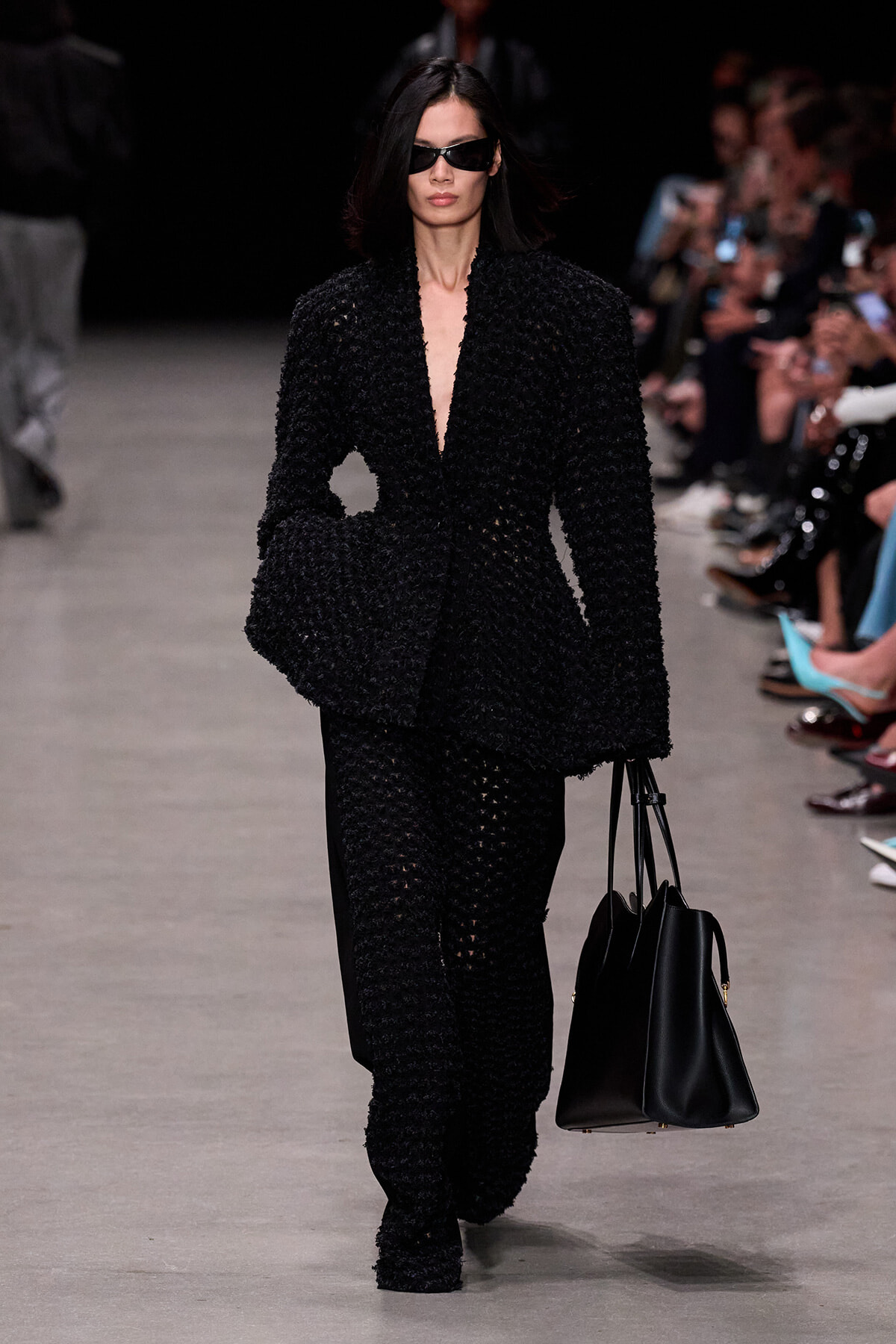 Model walks the runway in an all-black textured suit with oversized jacket, black sunglasses, and a black tote bag.