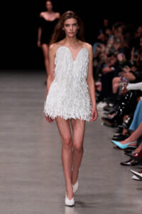 Model wearing a white fringed strapless mini dress walks the runway, audience watching in the background.