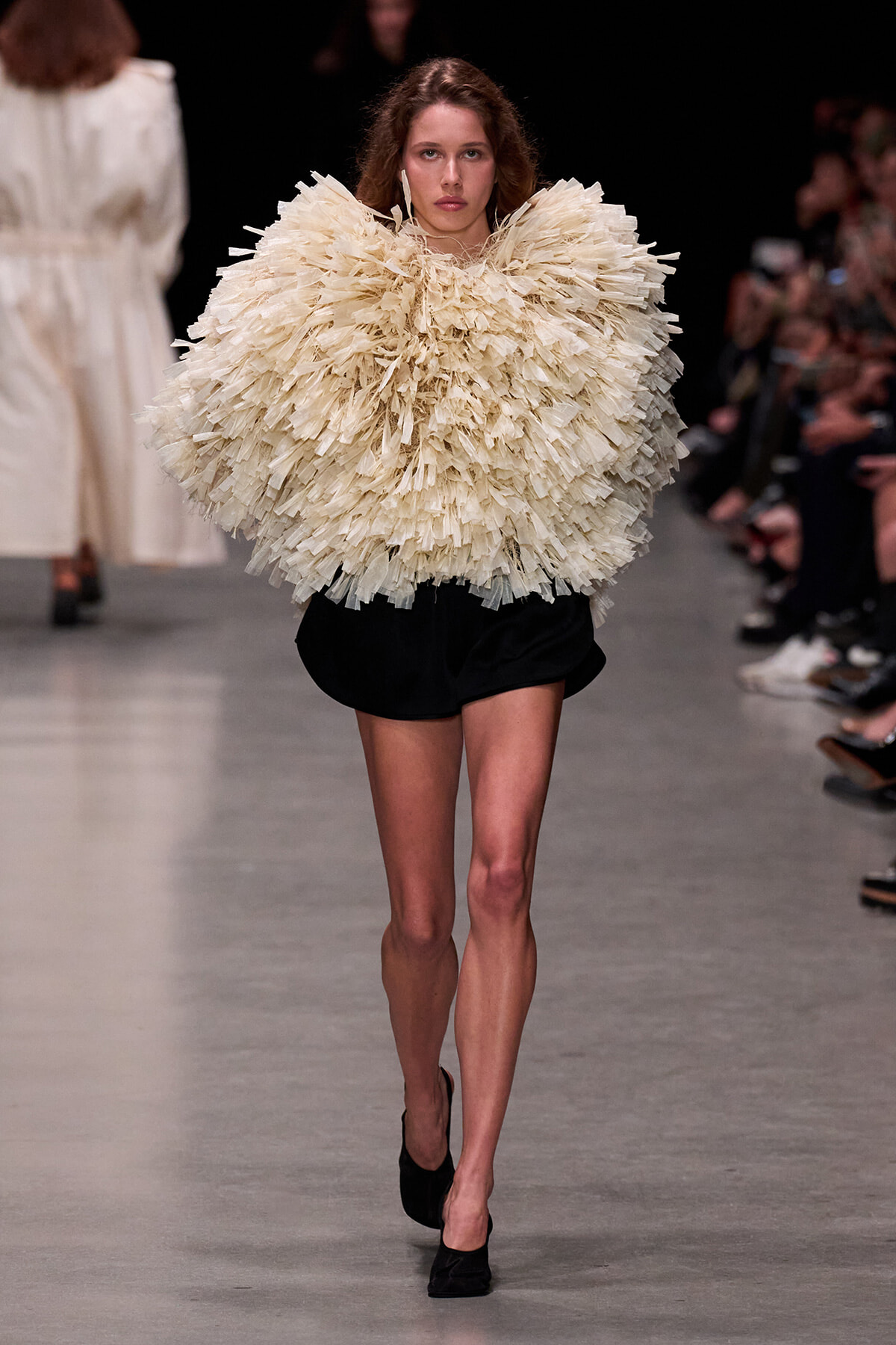 Model walking the runway in a voluminous cream feathered top and black shorts; bold fashion statement on a catwalk.