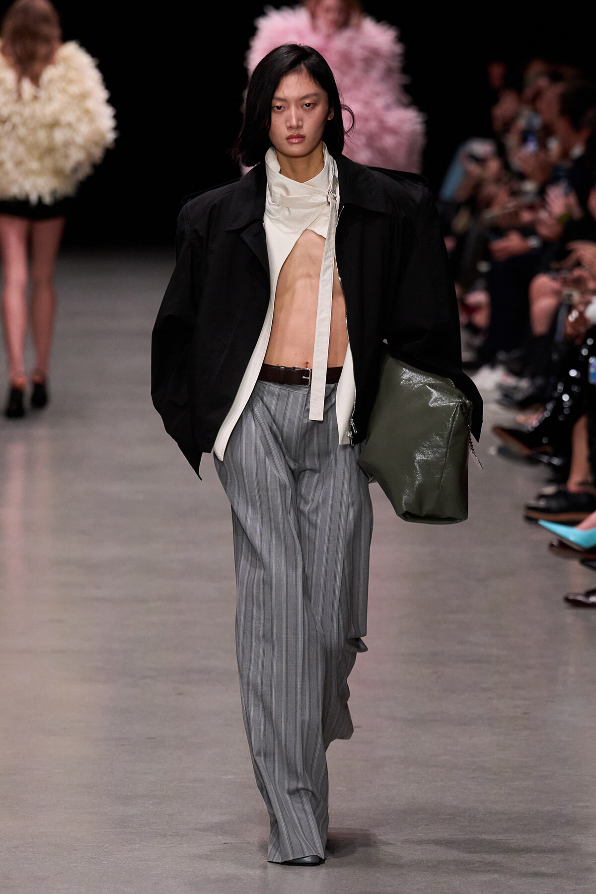 Model walks the runway in a black oversized jacket over a cream top, exposing midriff, paired with gray pinstripe wide-leg pants and a brown belt, carrying a large olive tote.