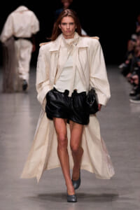 Model on runway wearing a beige oversized trench coat over a light top and black leather shorts, gray heels, walking confidently down the catwalk.