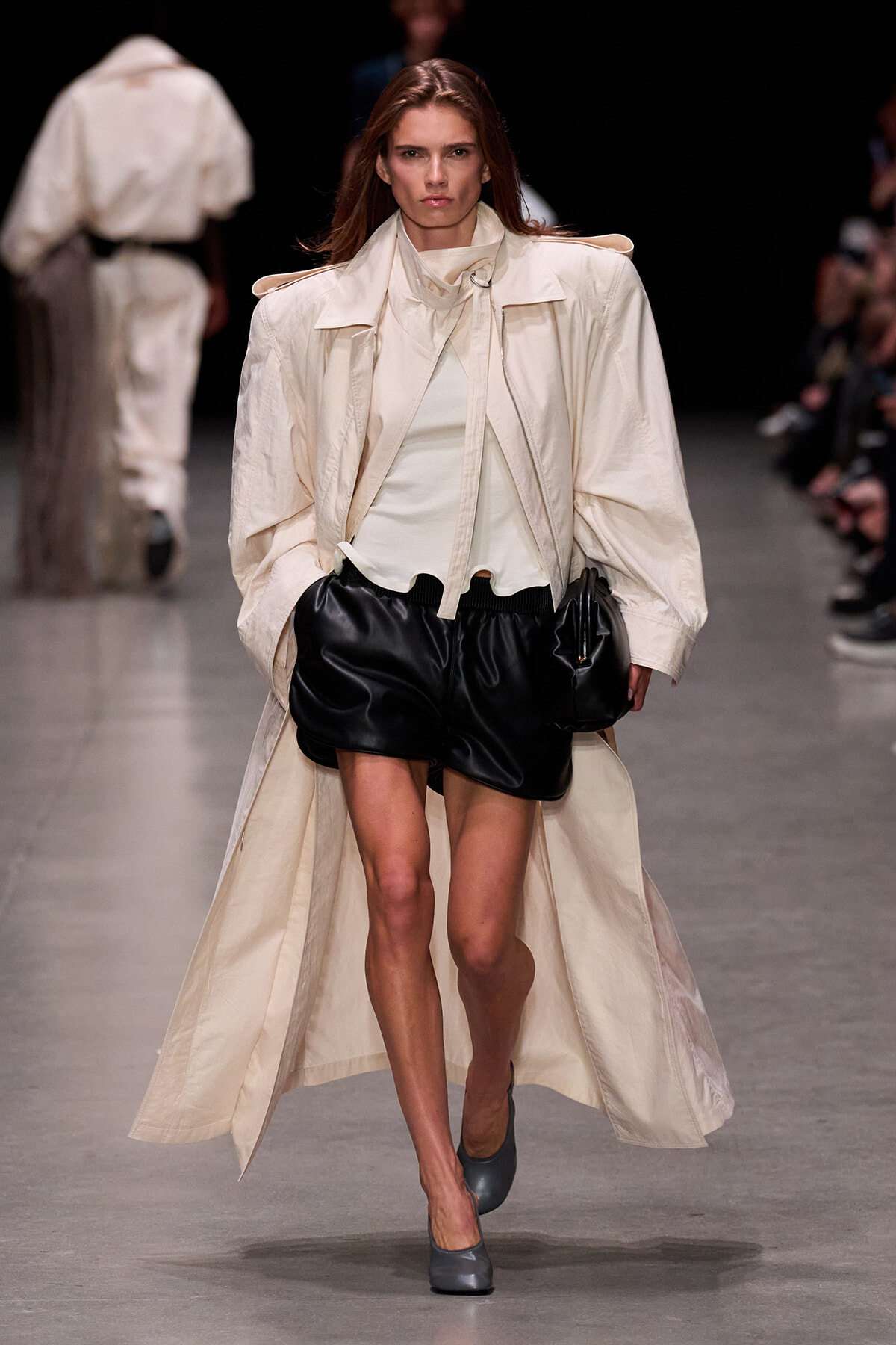 Model on runway wearing a beige oversized trench coat over a light top and black leather shorts, gray heels, walking confidently down the catwalk.