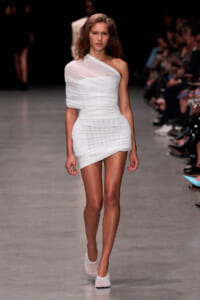 Model walks the runway in a white one-shoulder, ruched mini dress with matching white heels.