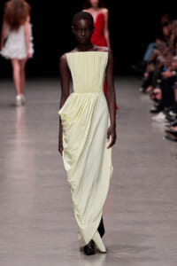 Model walks the runway in a pale yellow, strapless pleated gown with a draped, gathered skirt and dark boots. Behind her, other models and audience blur in the background.