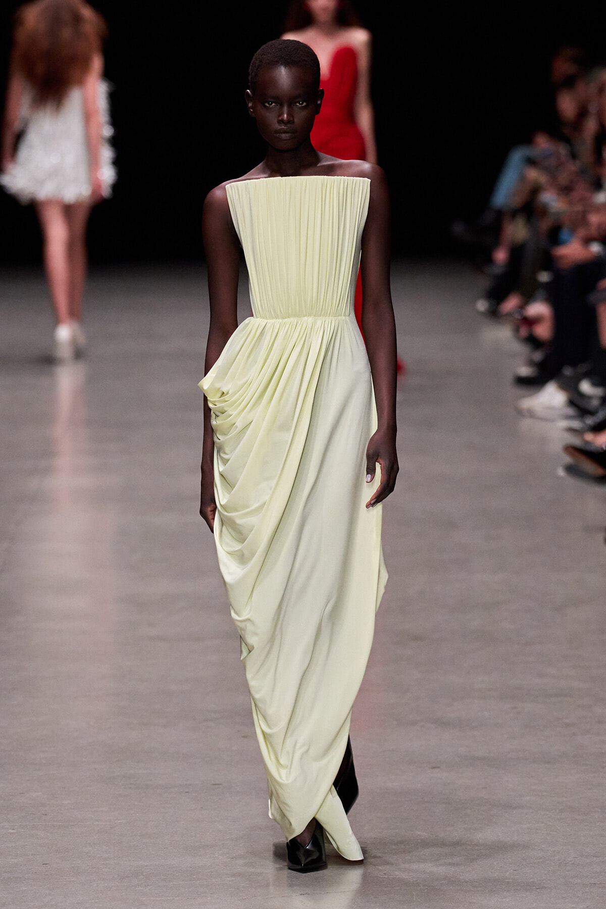 Model walks the runway in a pale yellow, strapless pleated gown with a draped, gathered skirt and dark boots. Behind her, other models and audience blur in the background.