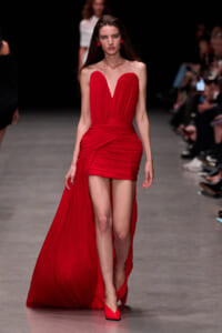 Model walks a runway in a red strapless gown with a sweetheart neckline and draped bodice, the dress knotting at the waist and a long train trailing behind her to the floor edge on the left.