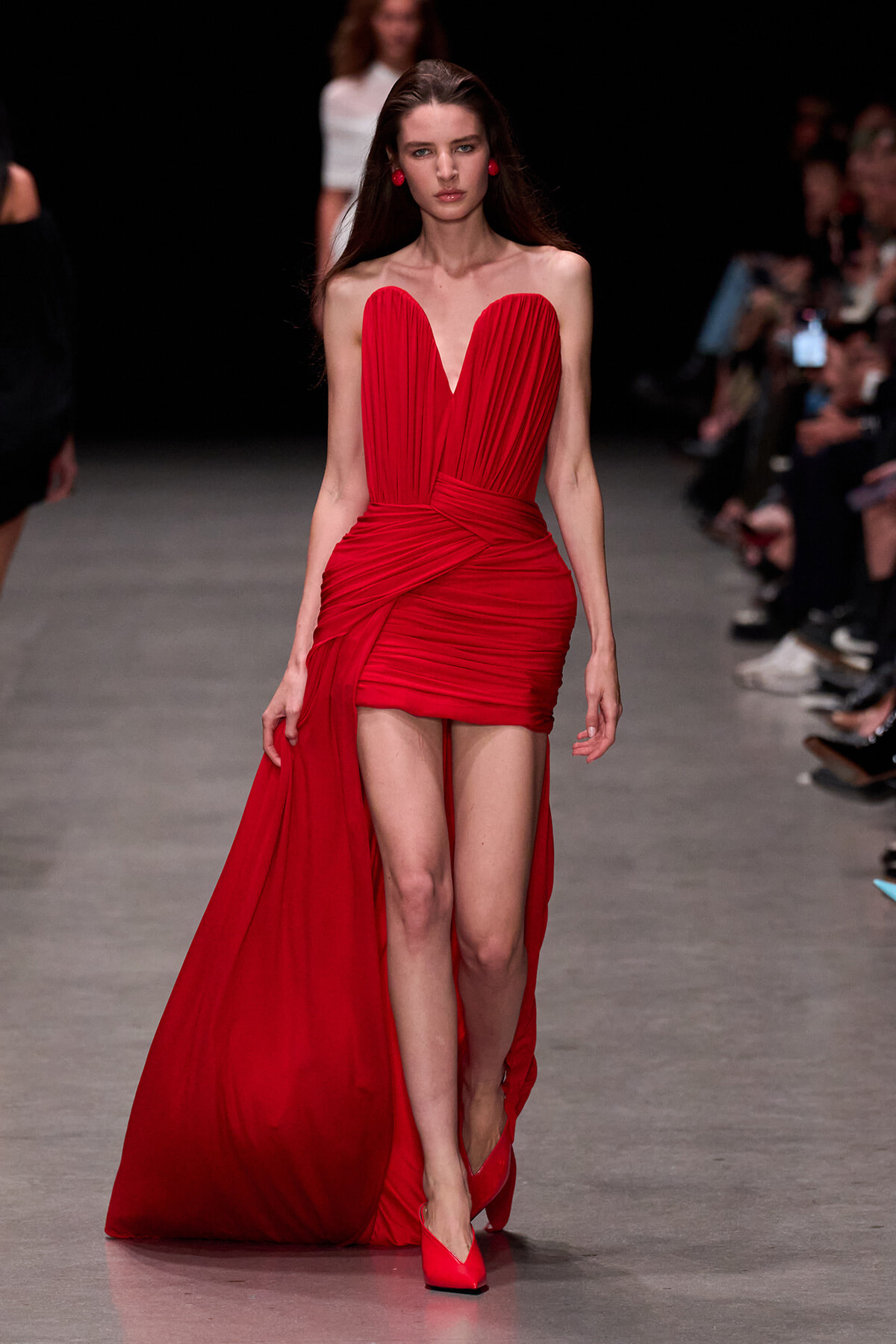 Model walks a runway in a red strapless gown with a sweetheart neckline and draped bodice, the dress knotting at the waist and a long train trailing behind her to the floor edge on the left.