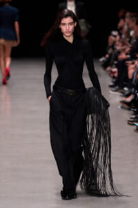 Model walking the runway in an all-black outfit: fitted, ruched long-sleeve top with wide-leg pants, carrying a long black fringe accessory.