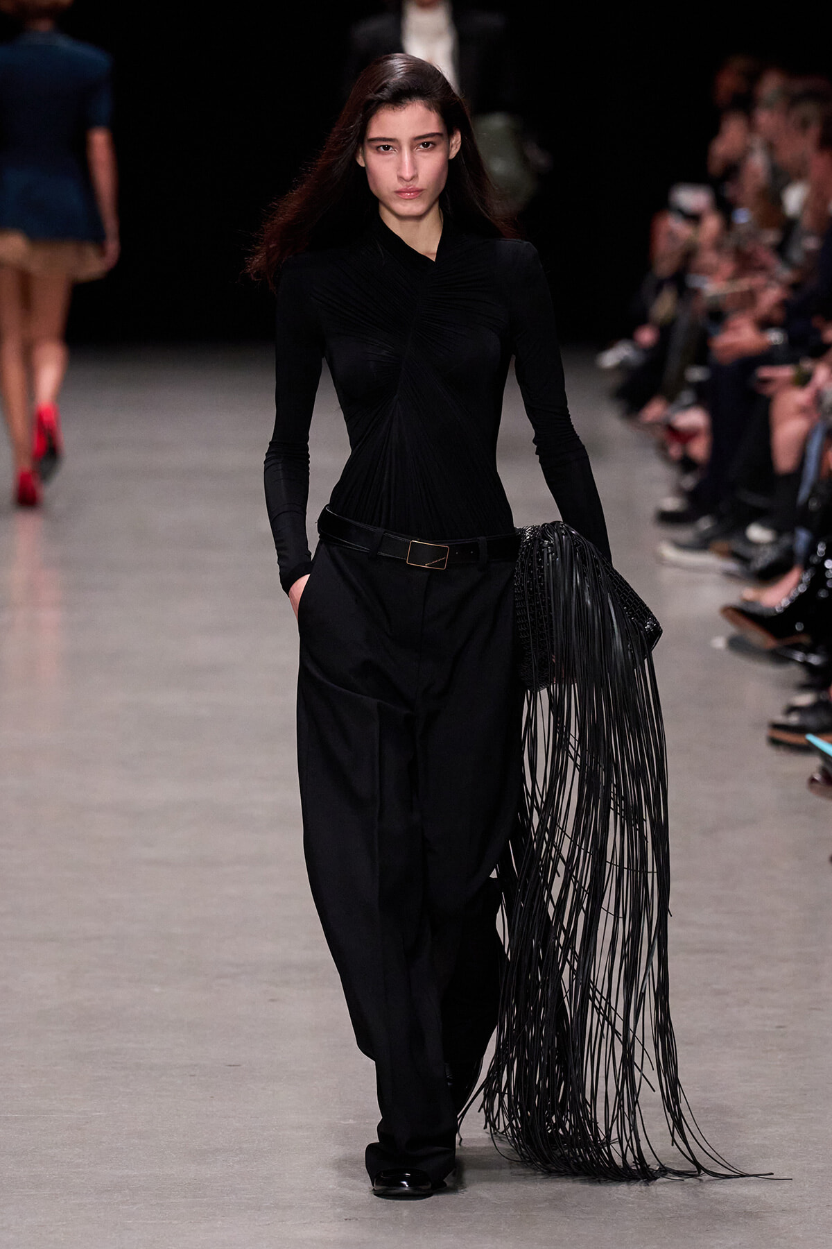 Model walking the runway in an all-black outfit: fitted, ruched long-sleeve top with wide-leg pants, carrying a long black fringe accessory.