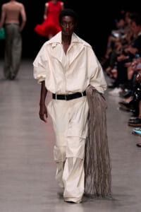 Black female model walks runway in oversized cream jumpsuit with a black belt, carrying a long fringe bag.