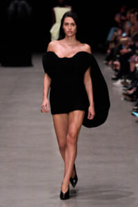 Model walking the runway in a black off-the-shoulder dress with a draped cape, paired with black heels and minimal accessories.