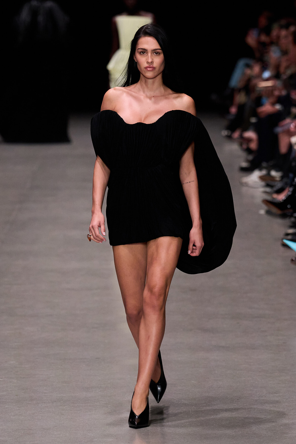 Model walking the runway in a black off-the-shoulder dress with a draped cape, paired with black heels and minimal accessories.