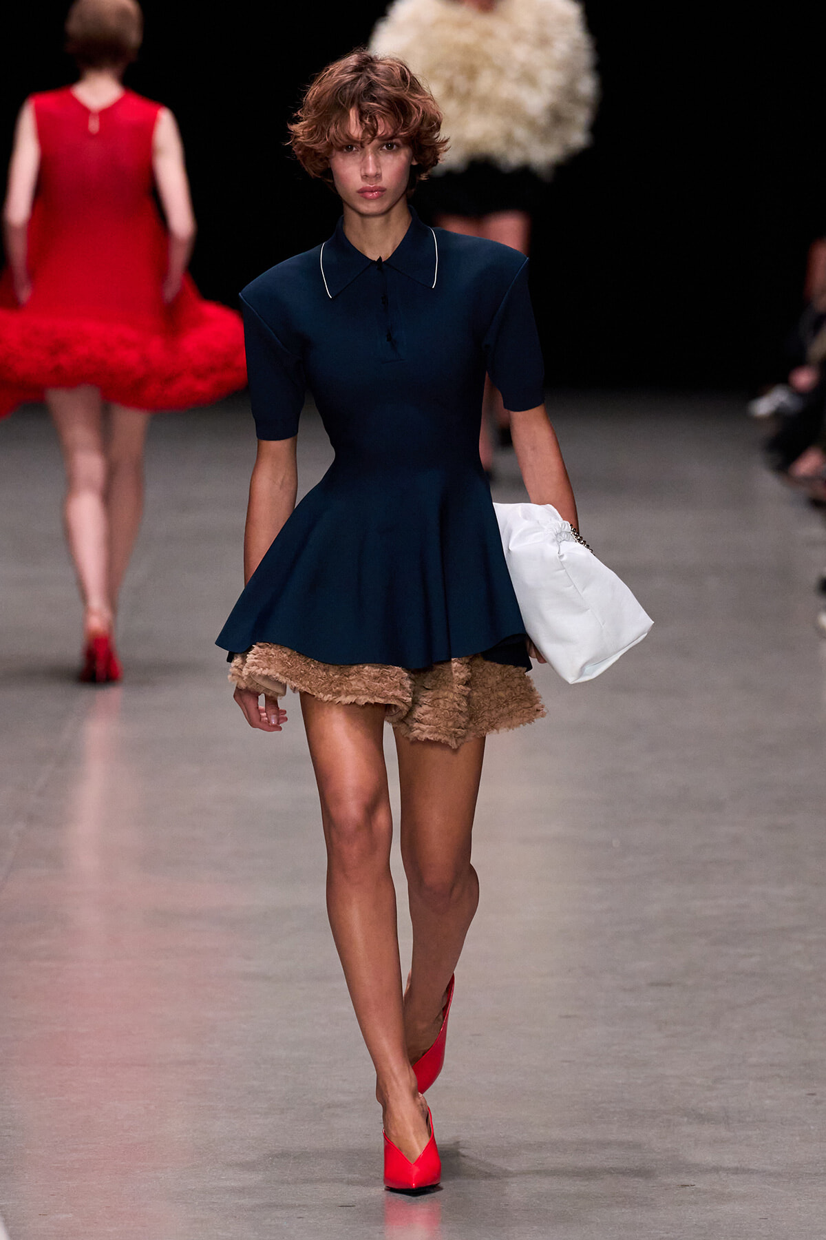 Model walking a runway in a navy short-sleeve dress with a peplum, beige textured shorts underneath, red heels, and a white clutch tote.