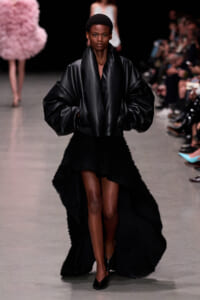 Model walks the runway in an oversized black leather jacket with voluminous sleeves and a high-low textured black skirt, black pumps.