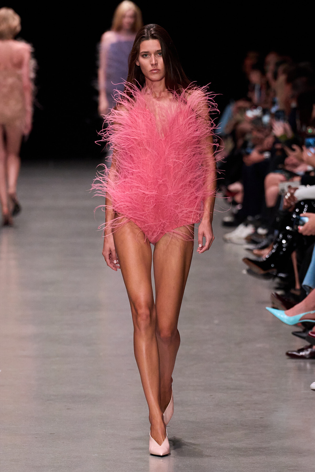 Model walking the runway in a pink feathered bodysuit paired with light pink heels, audience visible on both sides.