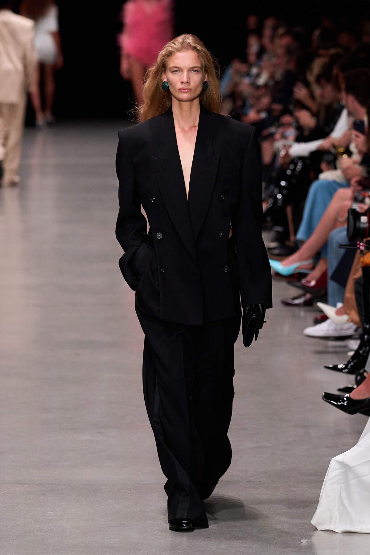 Model walking the runway in an oversized black double-breasted blazer with wide-leg pants and green statement earrings.