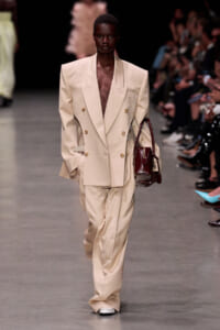 Model walks the runway in a cream oversized double-breasted suit with gold buttons, carrying a burgundy handbag.