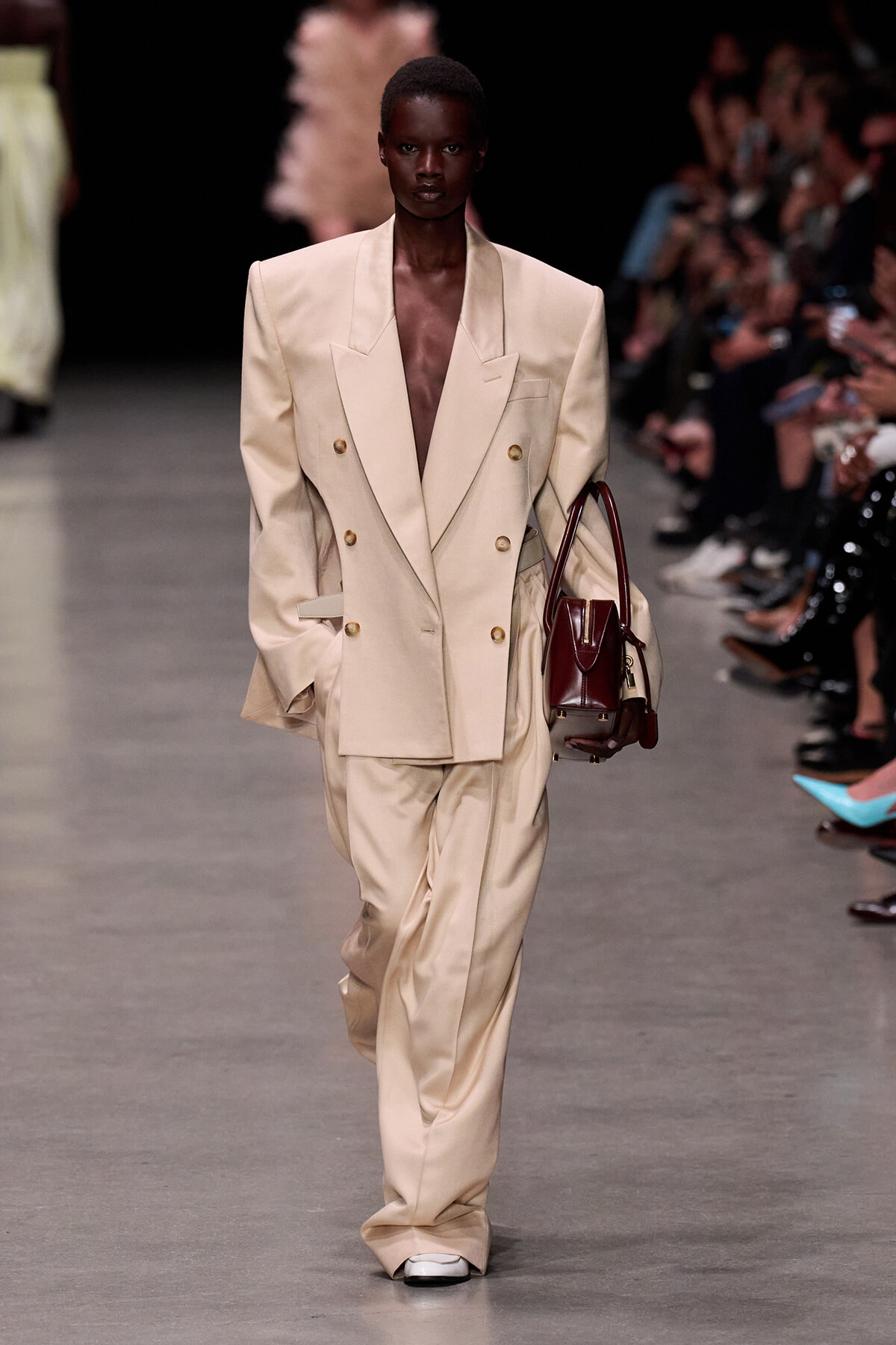 Model walks the runway in a cream oversized double-breasted suit with gold buttons, carrying a burgundy handbag.