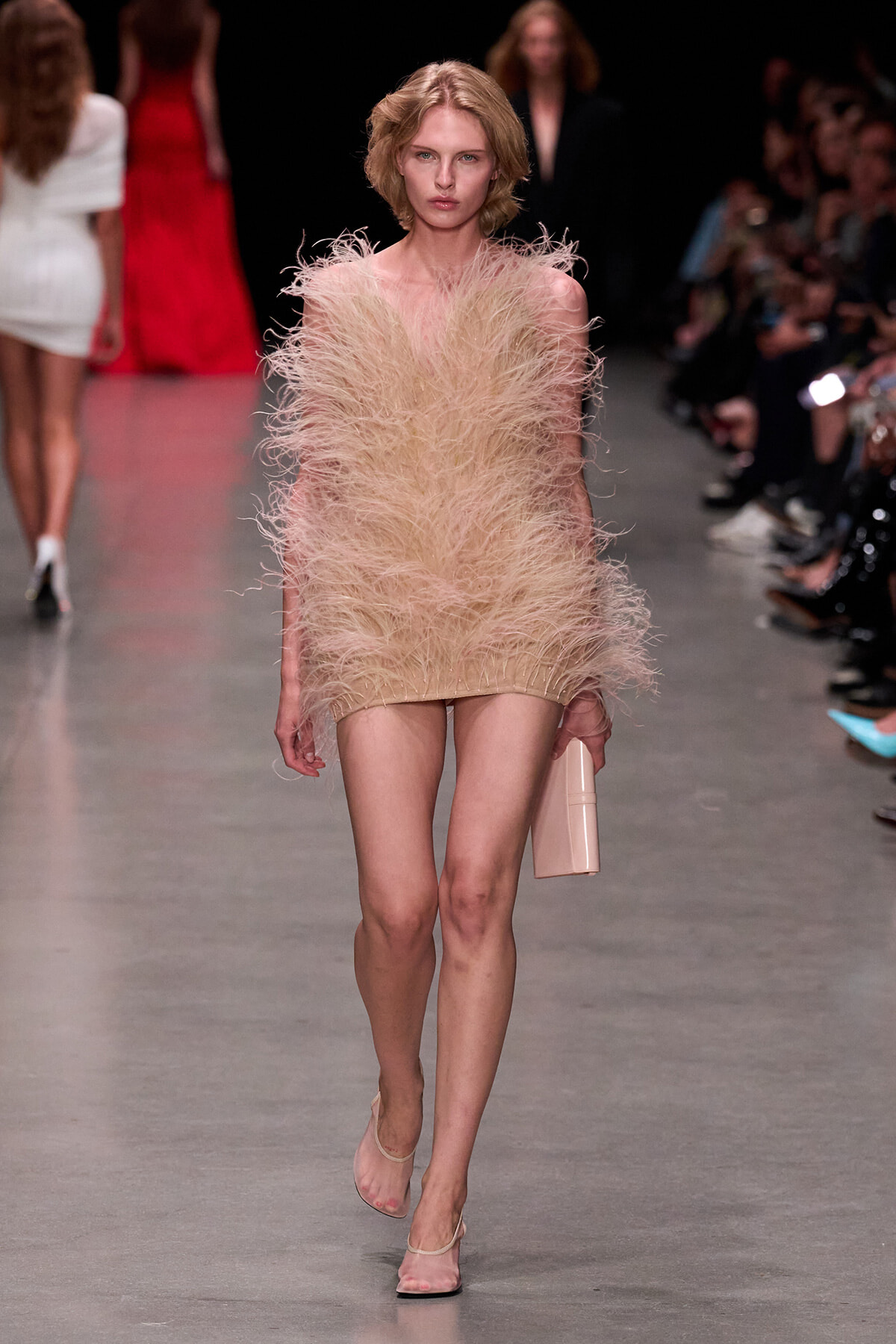 Model walking the runway in a beige feathered mini dress, nude sandals, and a pale pink clutch; soft, couture flair in a fashion show setting.