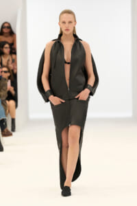 Model walking the runway in a black leather jumpsuit with cape sleeves and a front zipper, paired with black flats.