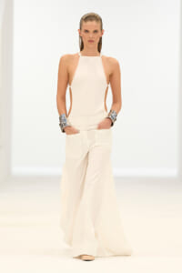 Model walking a runway in a white halter top with side cutouts and wide-leg white pants, paired with chunky silver cuffs on each wrist.