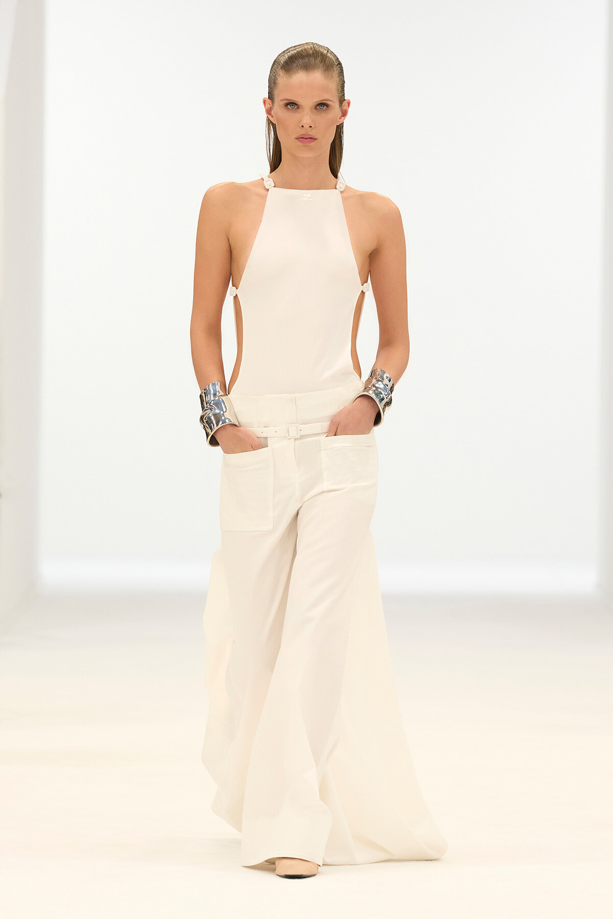 Model walking a runway in a white halter top with side cutouts and wide-leg white pants, paired with chunky silver cuffs on each wrist.