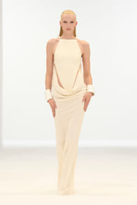 Model on a white runway wearing a cream halter-neck draped jumpsuit with wide cuff bracelets.