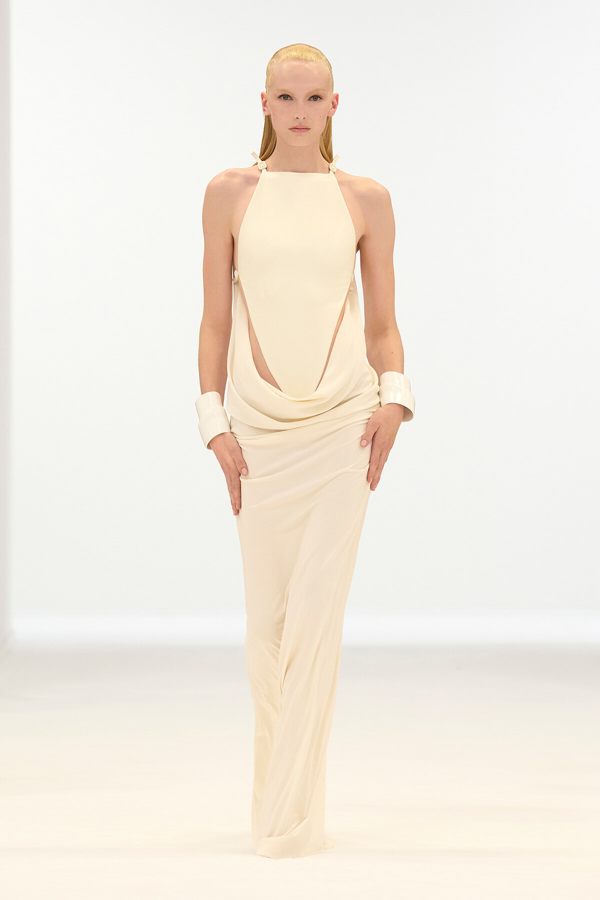 Model on a white runway wearing a cream halter-neck draped jumpsuit with wide cuff bracelets.