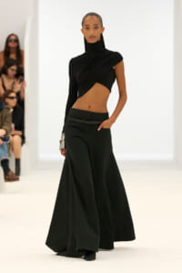 Model walking a fashion runway in a black one‑shoulder, cropped top and wide-leg pants, with a metallic bracelet visible on her left wrist.