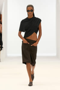 Model on a white runway wearing a black draped crop top, dark knee-length shorts, and black sunglasses.