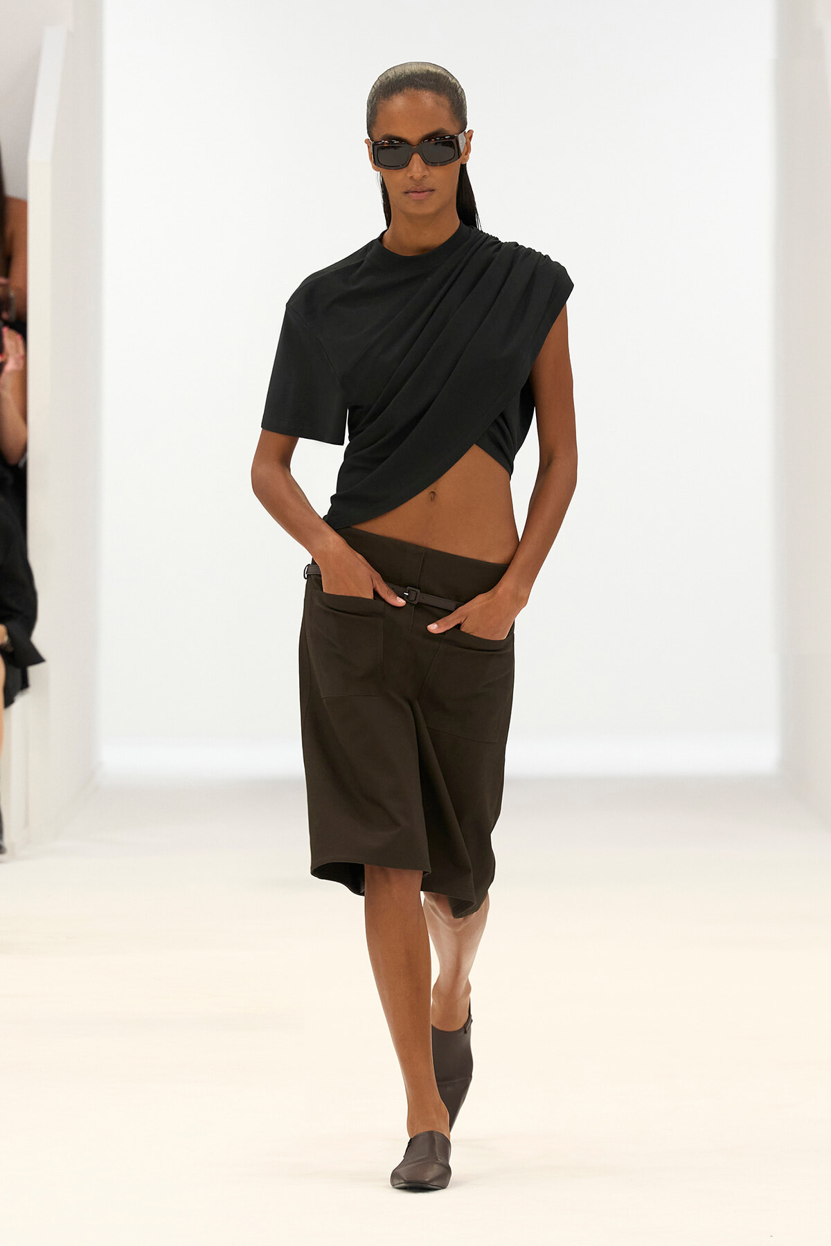Model on a white runway wearing a black draped crop top, dark knee-length shorts, and black sunglasses.