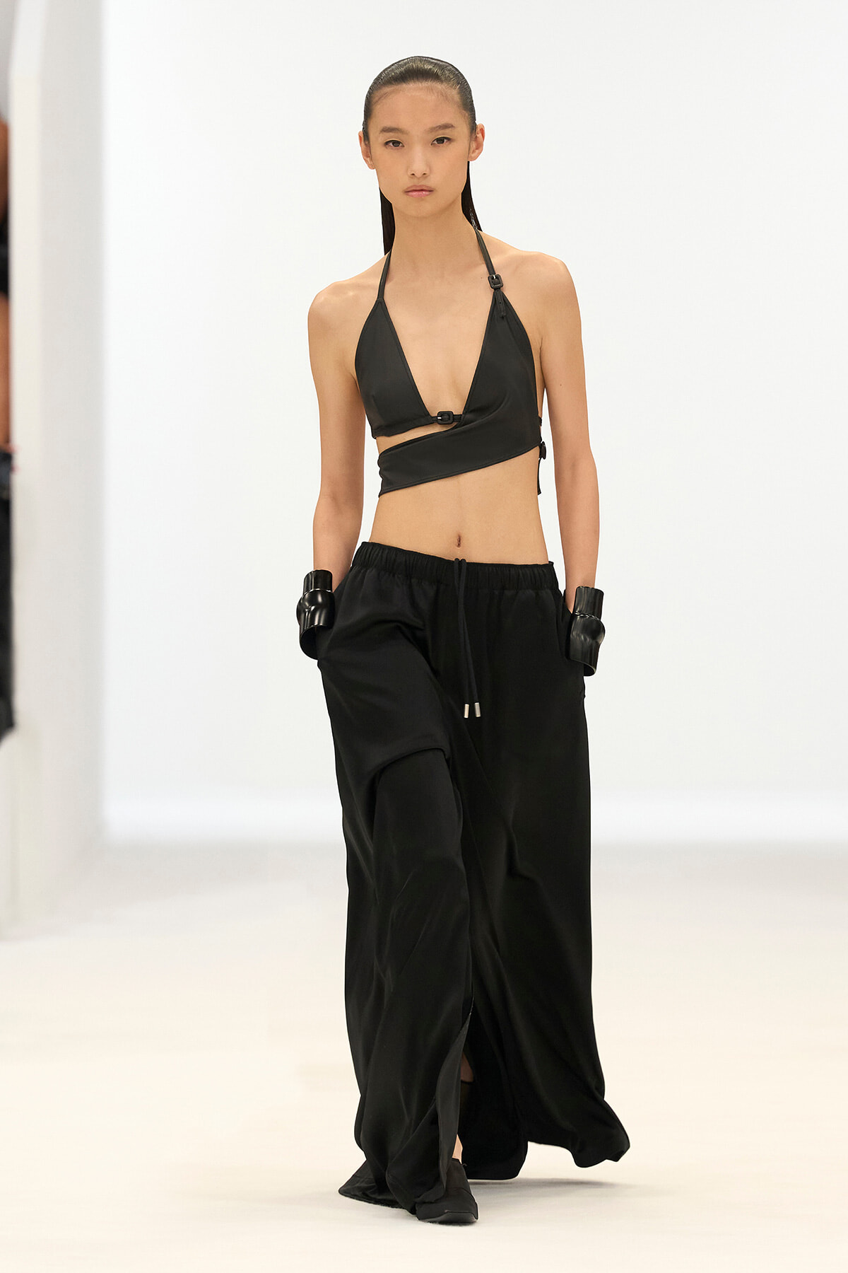 Model walking the runway in a black halter bikini top and loose black pants, wearing black gloves, against a white minimalist backdrop.