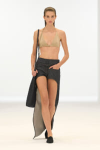 Model walking a runway in a beige triangle bikini top, dark denim skirt with long slits, and a shoulder bag slung over the shoulder.