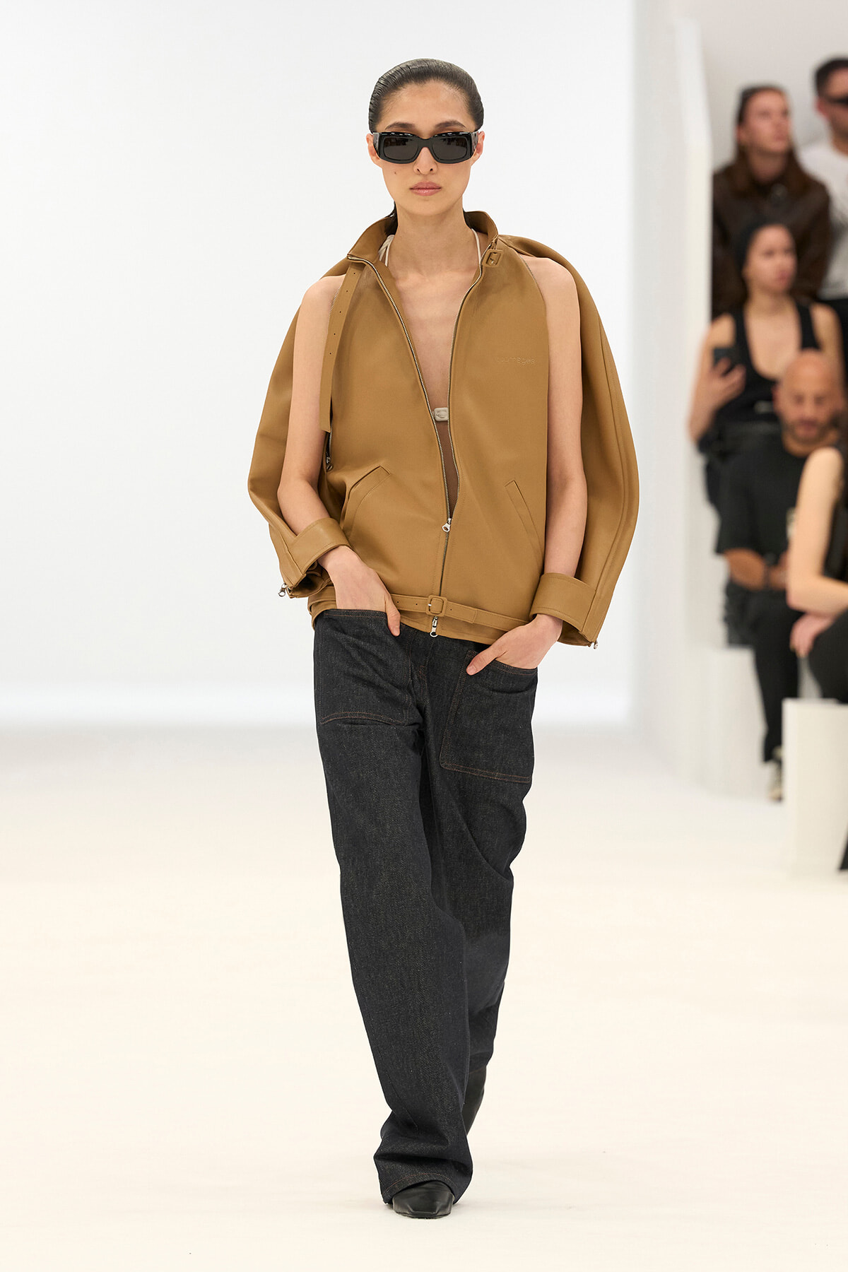 Model walking runway in oversized camel leather jacket with zipper, dark wide-leg jeans, and black sunglasses, hands in pockets.