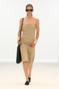 Model wearing a tan strapless midi jumpsuit with a thin belt, black shoulder bag, and black sunglasses on a white backdrop.