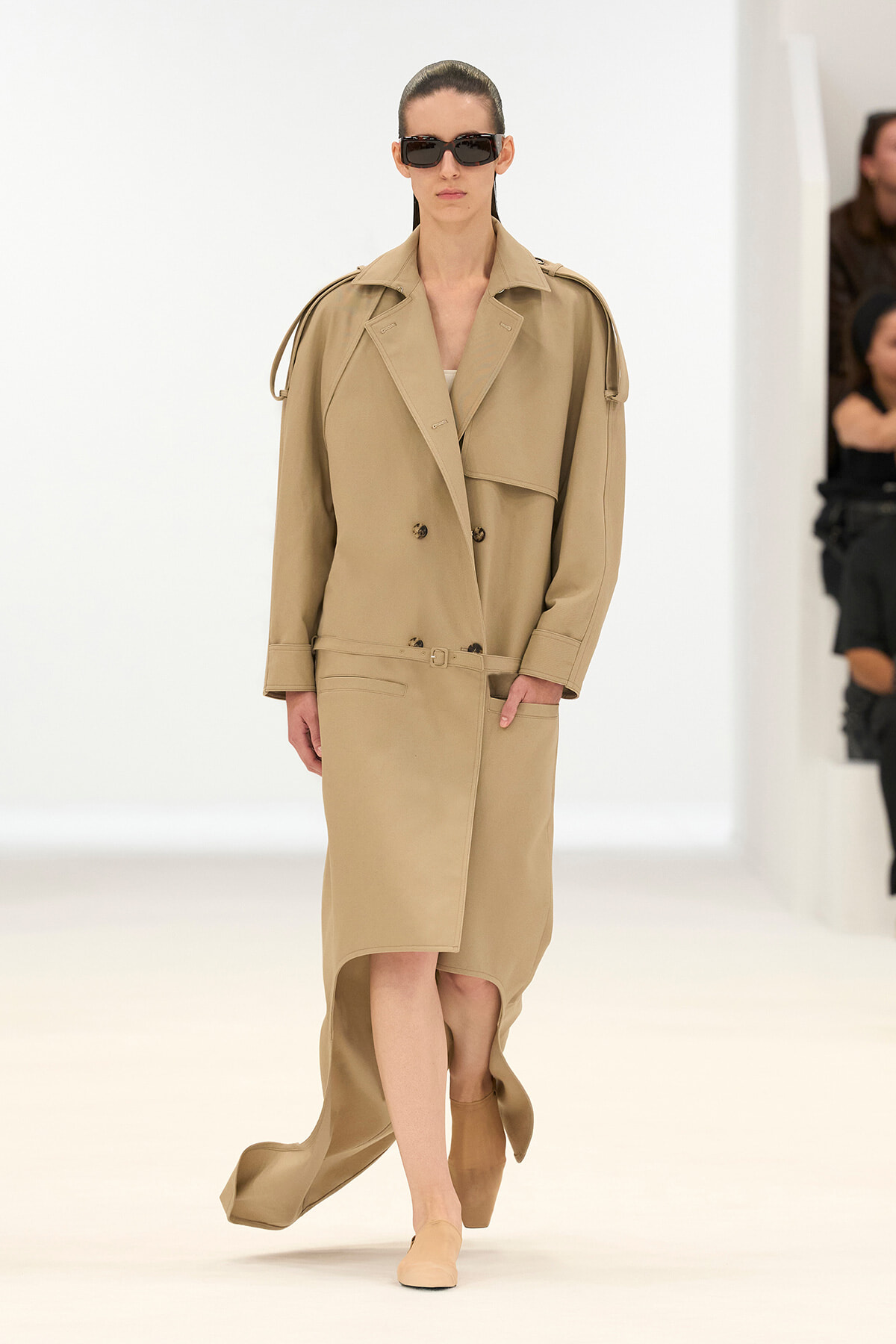 Model walks runway in a beige oversized trench coat with belt, holding a neutral pose and wearing dark sunglasses.
