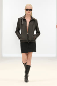 Model walking a runway in a black leather zip-front jacket, matching mini skirt, knee-high boots, and dark sunglasses.