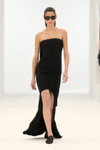 Model walking a runway in a strapless black gown with a thin belt and a high front slit, wearing dark sunglasses.