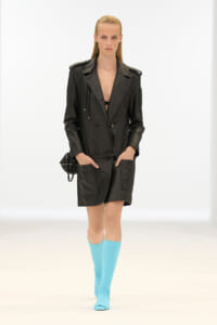 Model wearing a black leather trench coat with a belt and large pockets, paired with bright blue knee-high socks on a runway.