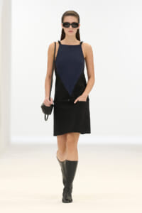 Model wearing a navy sleeveless top with a black triangular panel, paired with a black mini skirt, knee-high boots, sunglasses, and a small crossbody bag.