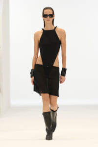Model walks a white runway in a black halter-neck dress with side cutouts, paired with black sunglasses, wrist cuffs, and knee-high boots.