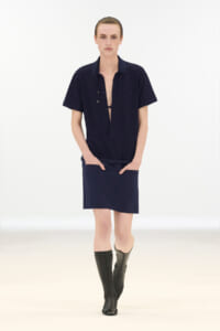 Model walking the runway in a navy short-sleeve shirt with open placket, matching navy skirt, and knee-high black boots.