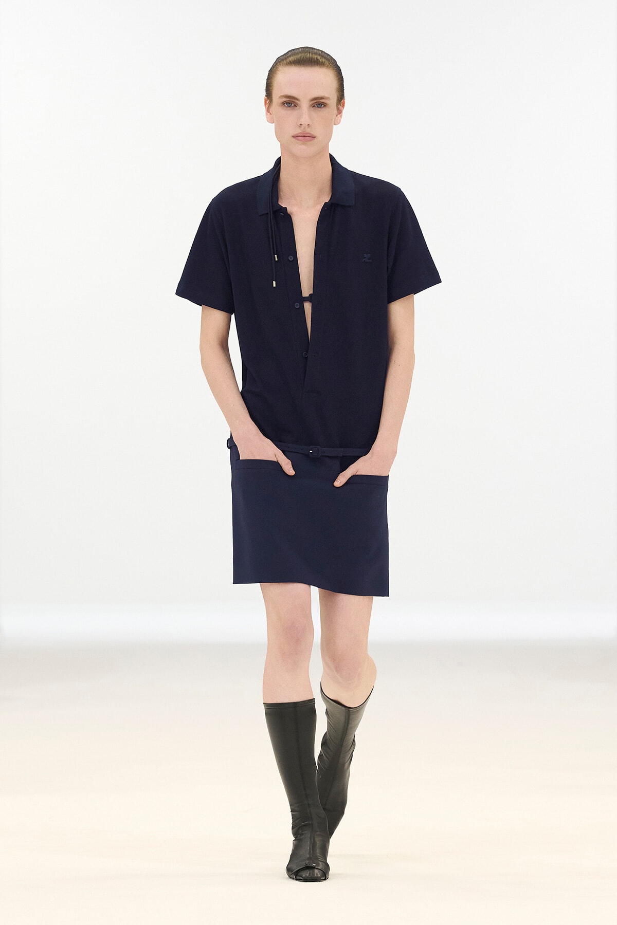 Model walking the runway in a navy short-sleeve shirt with open placket, matching navy skirt, and knee-high black boots.